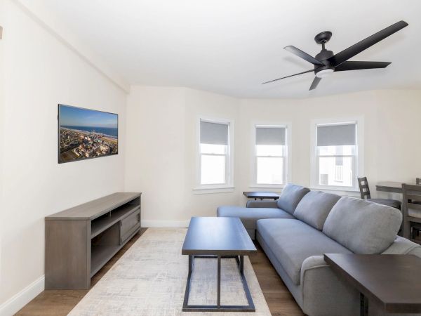 A modern living room features a gray sofa, ceiling fan, TV, coffee table, and dining area with light walls and three windows.